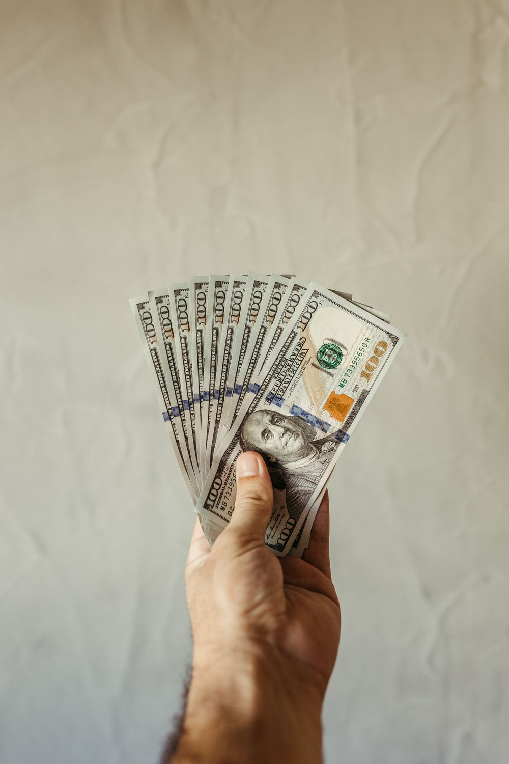 A hand displays a fan of US hundred dollar bills against a plain background.