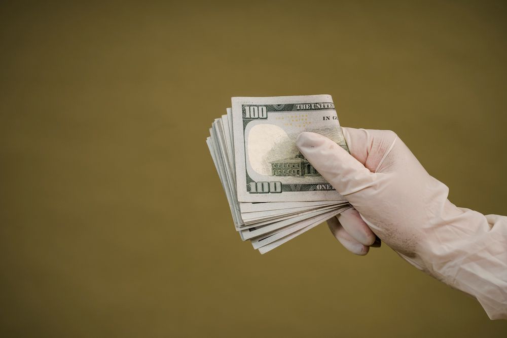 A gloved hand holds a stack of hundred-dollar bills against a plain background.
