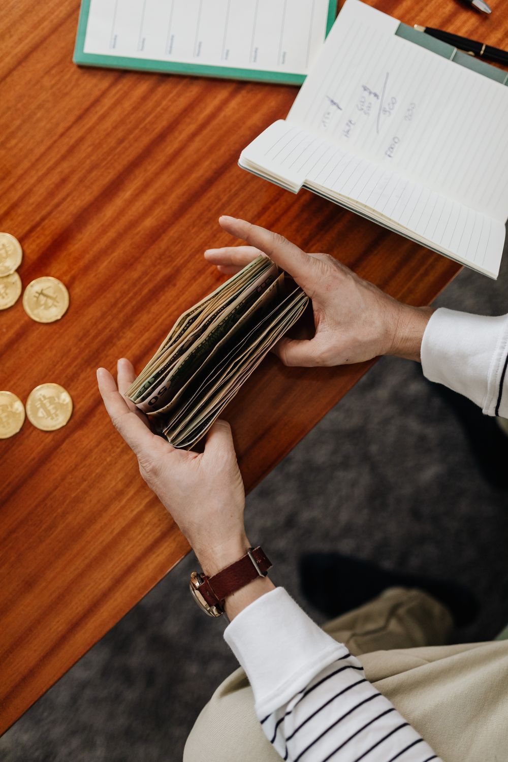 A person counting cash with coins and notebook on a wooden table, top view.