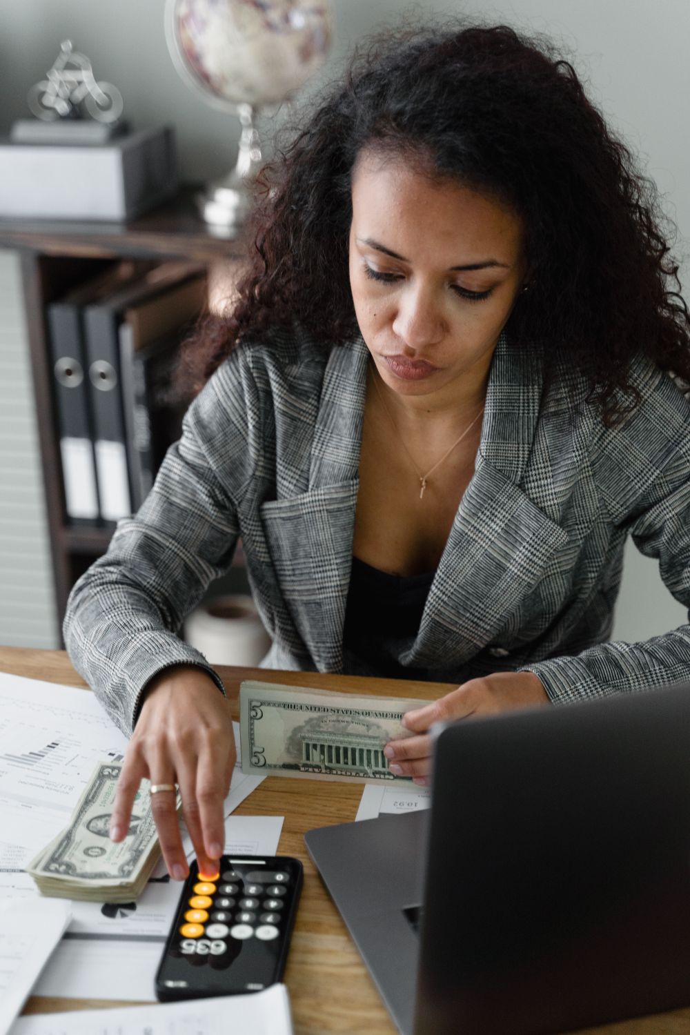 Focused businesswoman handling cash and calculations at her office desk, wearing a plaid blazer.
