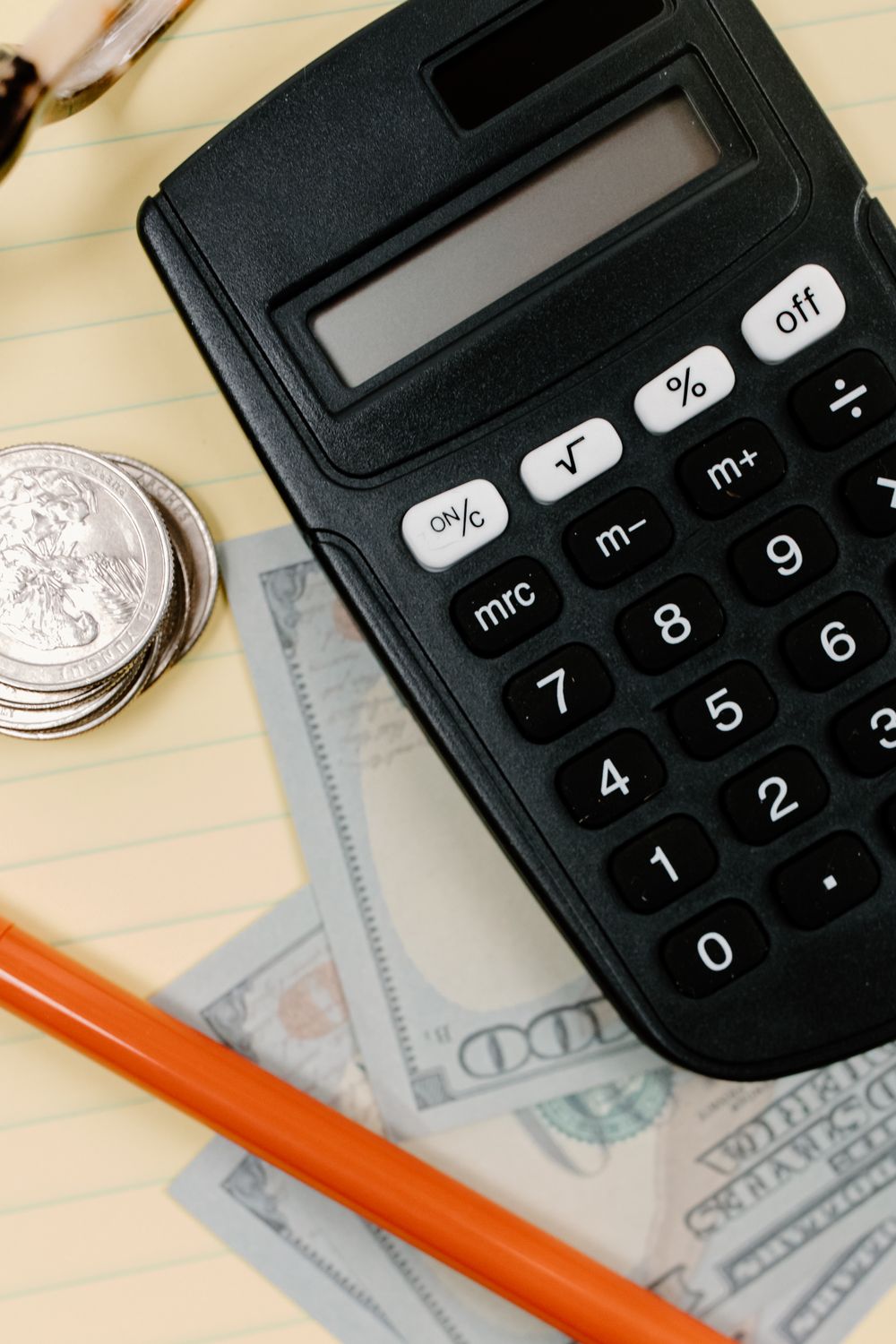 Black calculator, coins, and cash on a yellow notepad with a pencil.