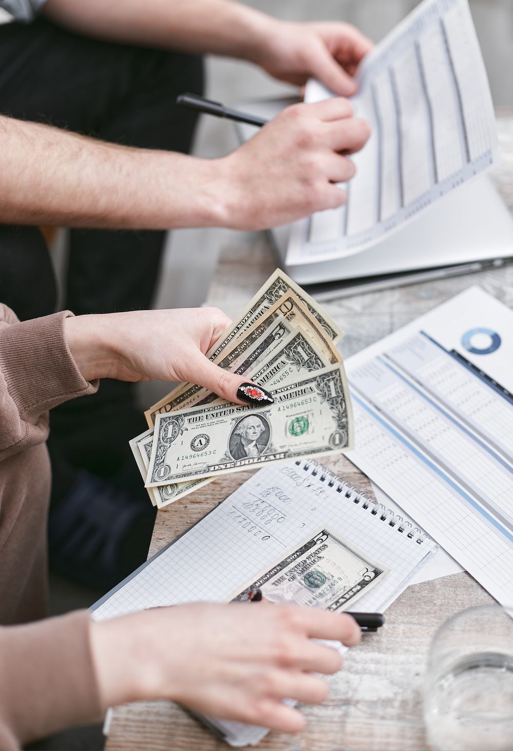 Two individuals counting dollar bills and reviewing financial documents indoors.